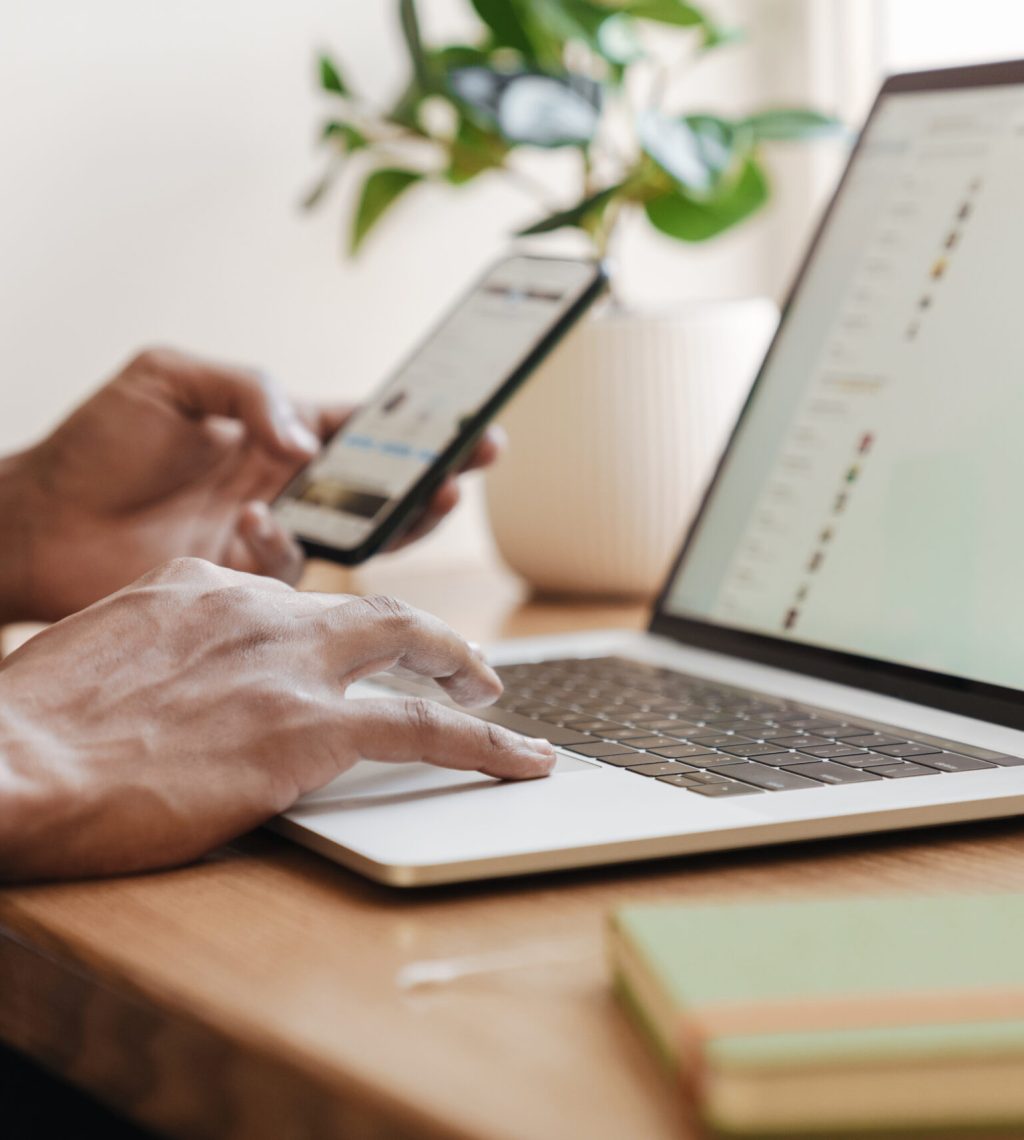 Cropped photo of young african guy indoors using laptop computer and mobile phone.
